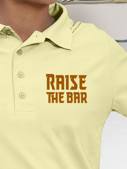 Close up of 'Raise The Bar' design on Women's Yellow Polo T-shirt, highlighting intricate details and vibrant print.